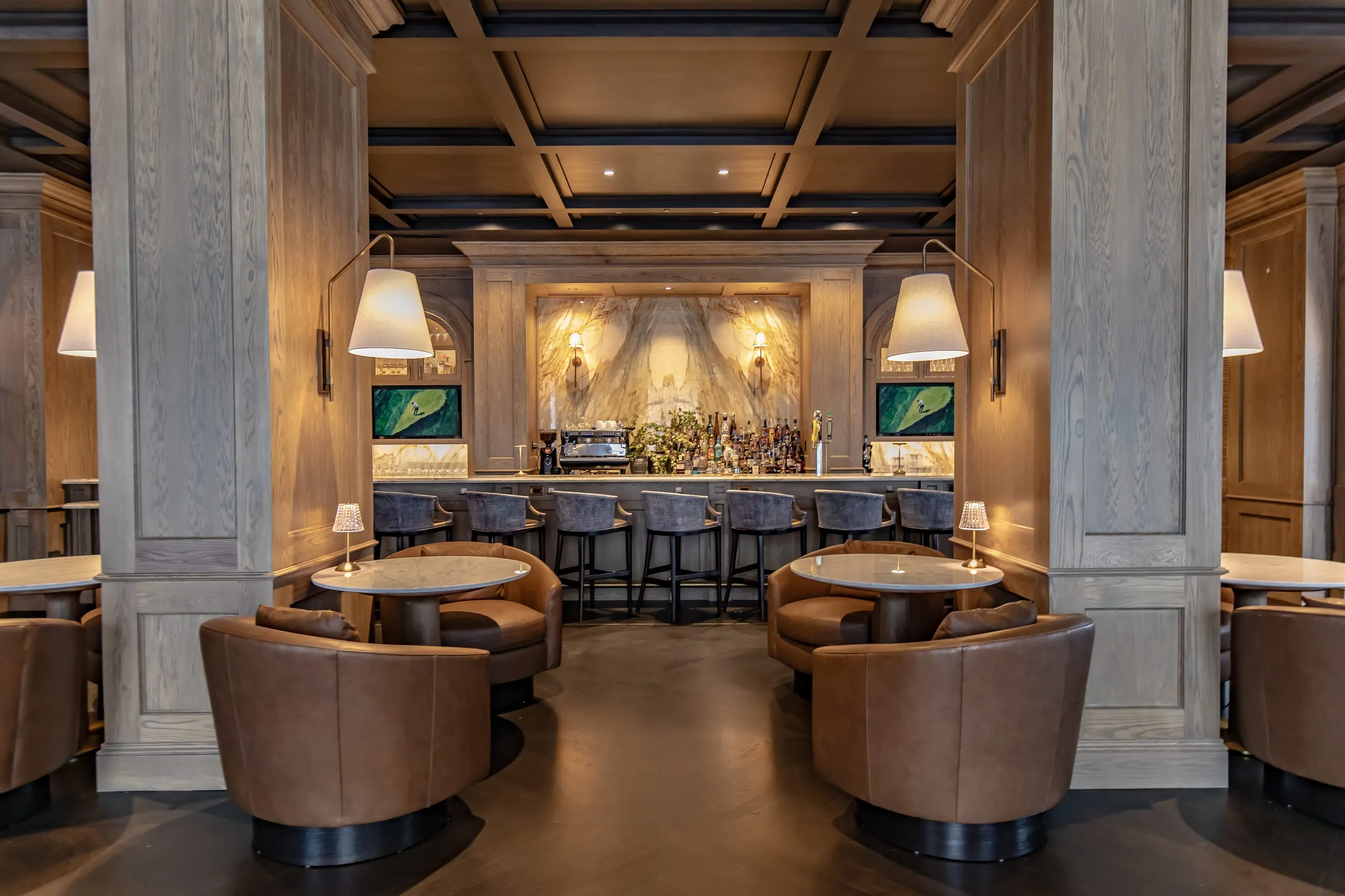 Club bar lounge with pale oak columns, tan leather swivel chairs at round tables and a backlit bar beyond