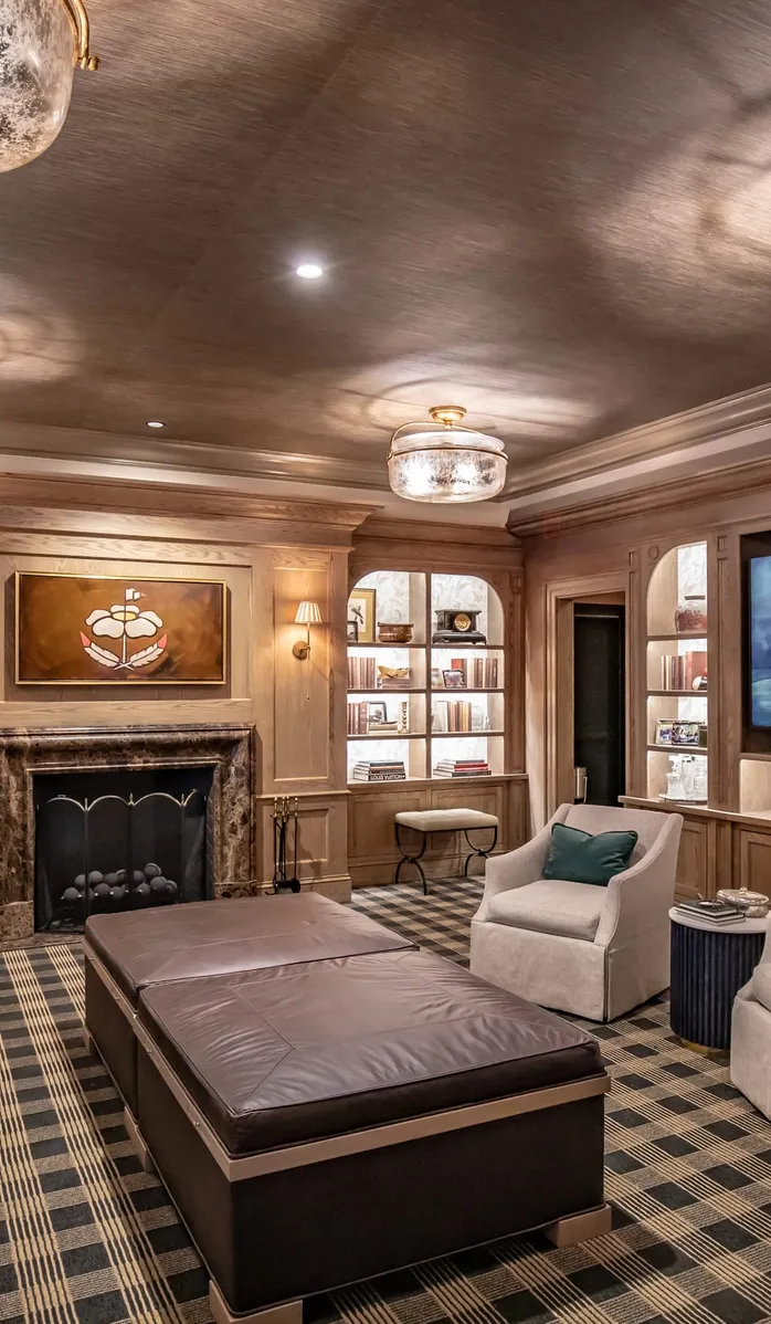 Private club lounge with a green velvet sofa, tufted leather ottoman and lit shelves flanking a fireplace on a plaid rug